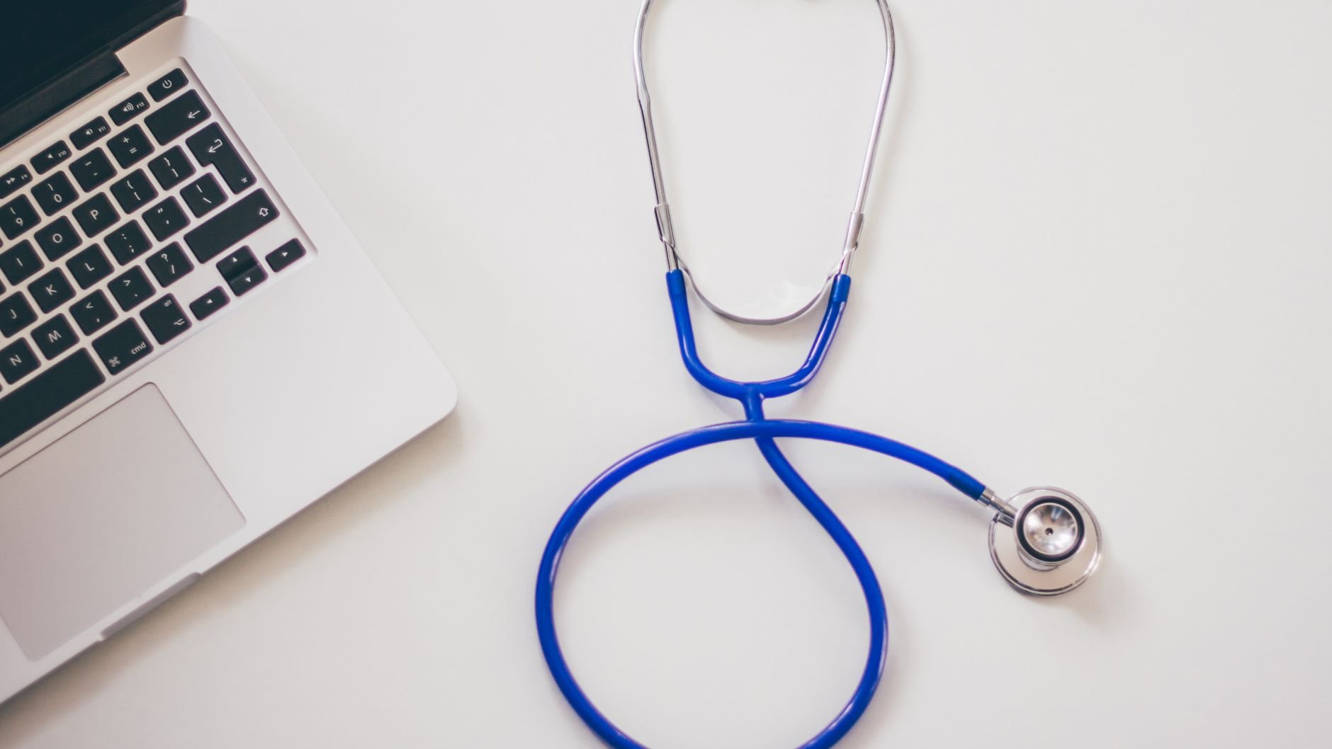 A blue stethoscope coiled on a white desk beside a laptop (illustrative stock photo)
