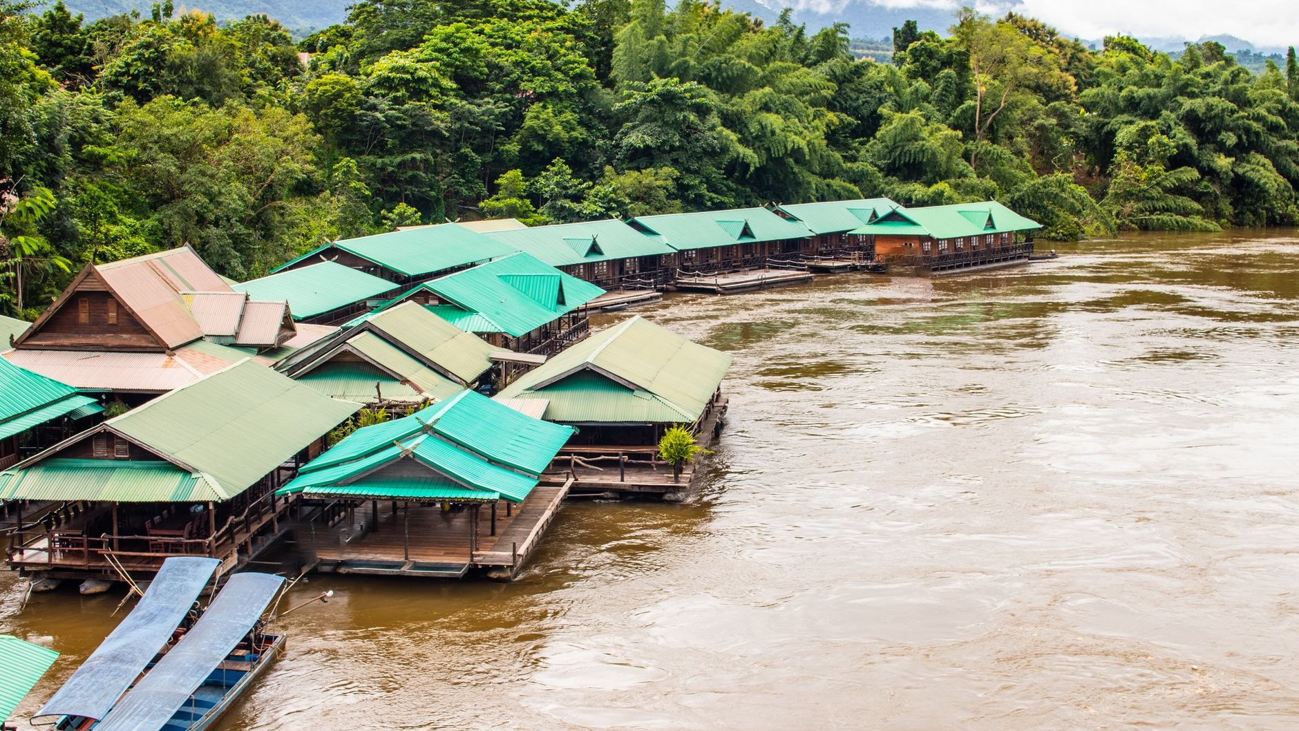 Green-roofed floating raft houses beside a brown river below forested hills in Kanchanaburi (illustrative stock photo)