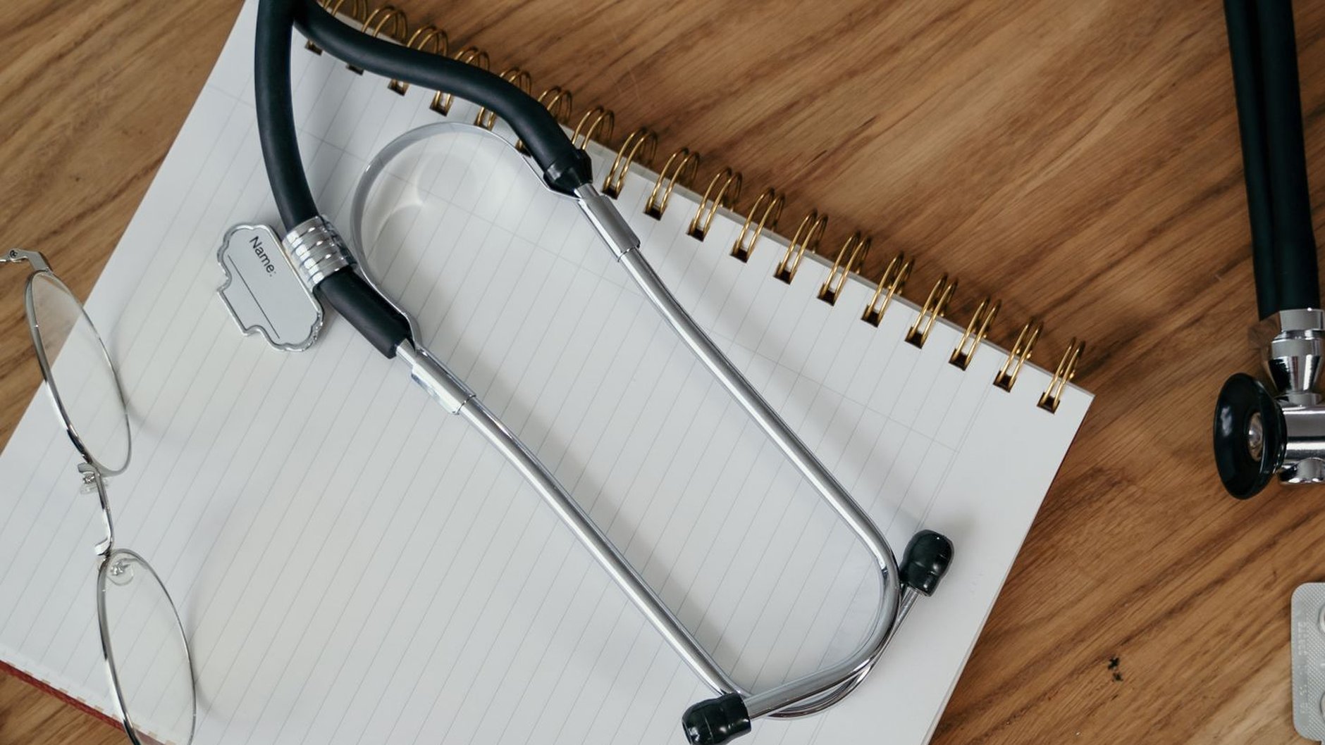 A stethoscope and a pair of glasses lie on a blank ruled notebook on a wooden desk (illustrative stock photo)