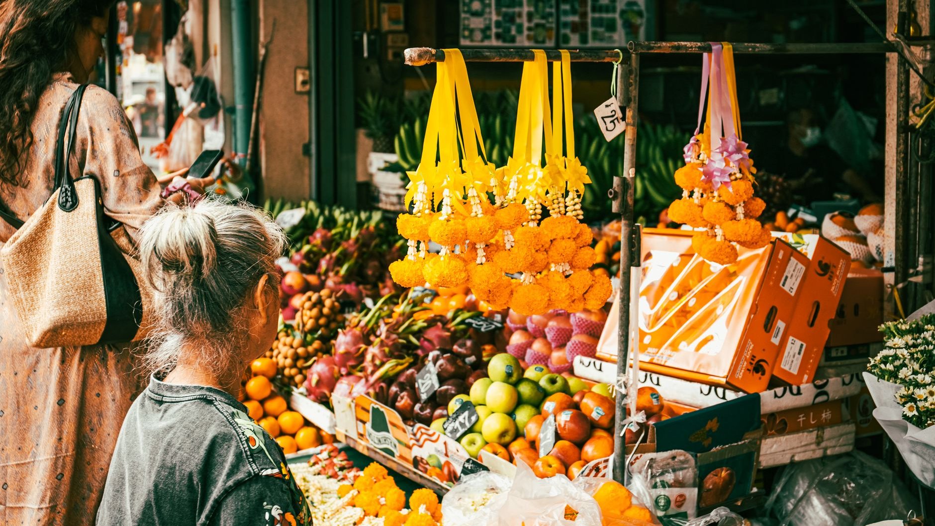 Two shoppers seen from behind at a Bangkok fruit stall, with garlands of marigolds hanging above dragon fruit, apples and oranges (illustrative stock photo)