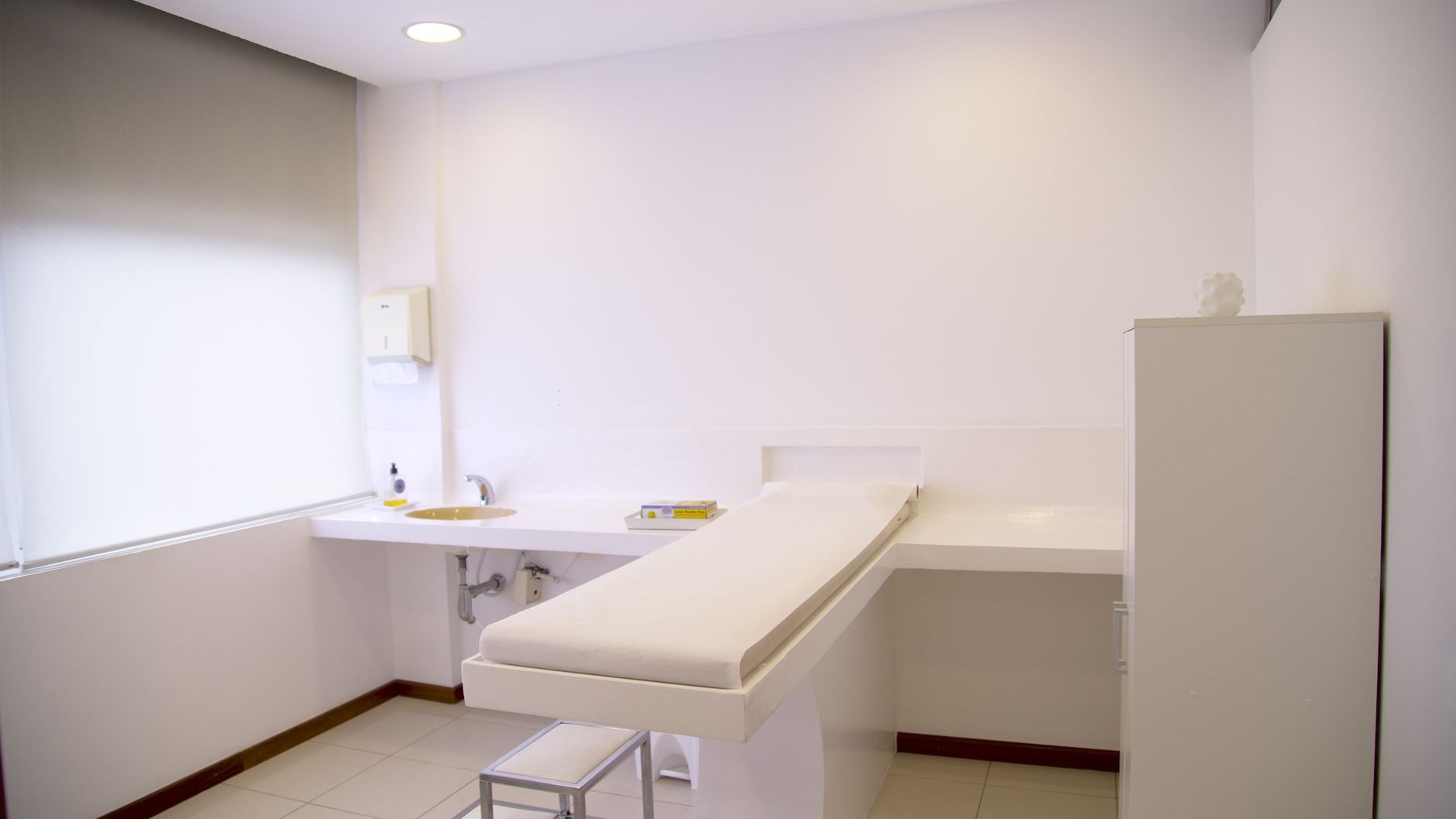 A bright, empty clinic room with a padded examination table and a washbasin (illustrative stock photo)