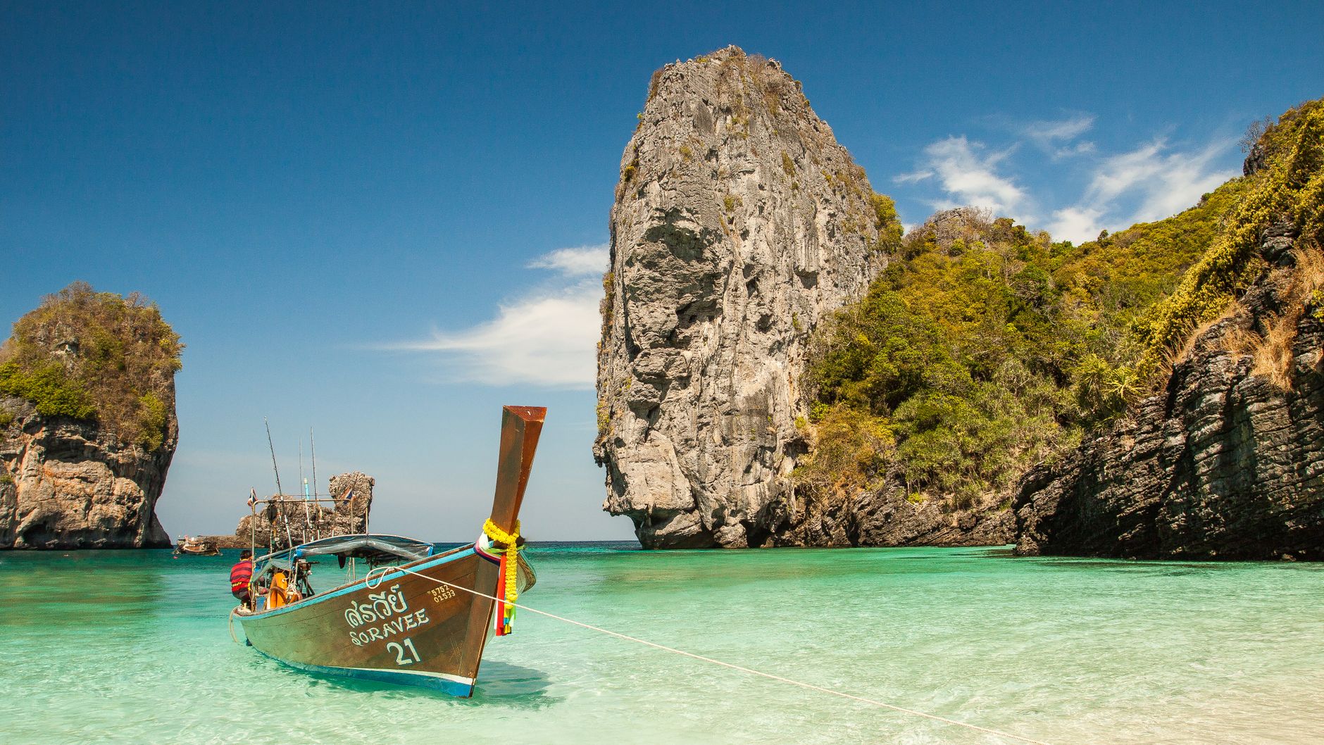 A longtail boat moored in clear shallow water beside limestone cliffs on Thailand's Andaman coast (illustrative stock photo)