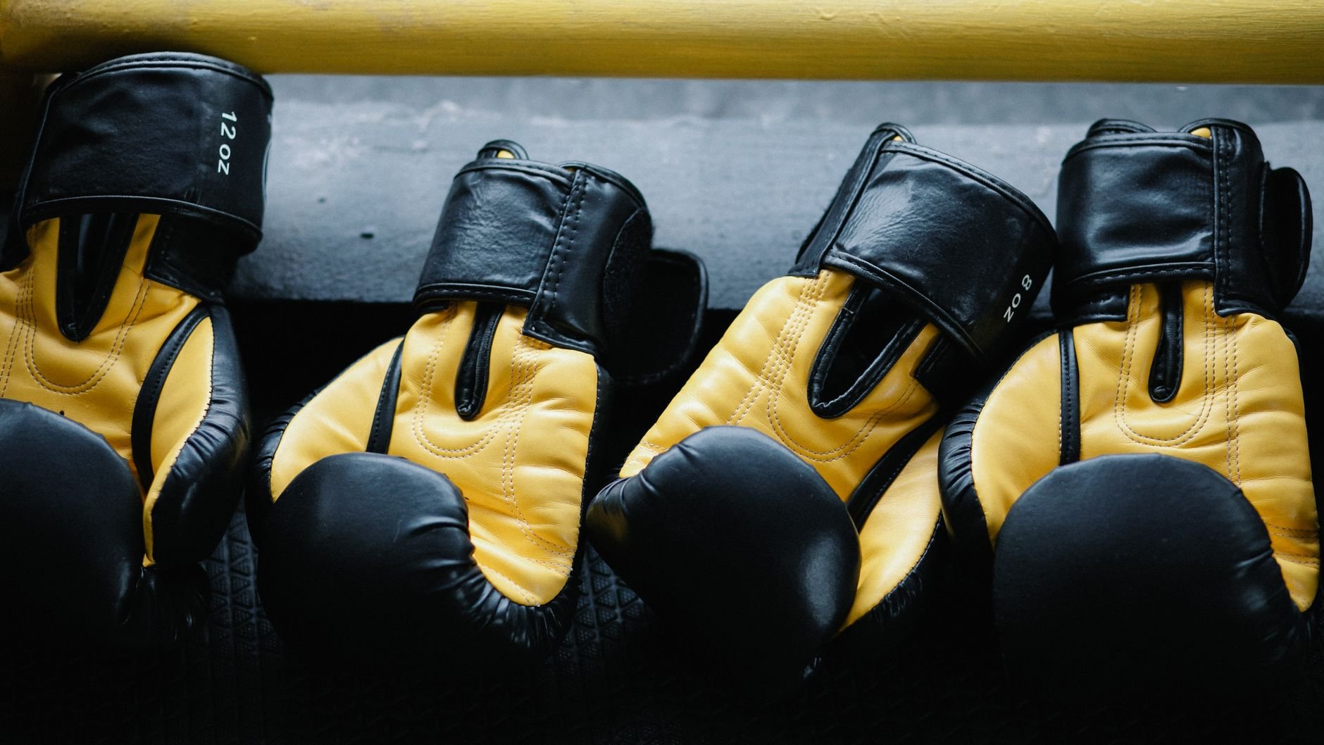 Four black and yellow 12 oz sparring gloves lined up on a ledge beside a yellow rail (illustrative stock photo)