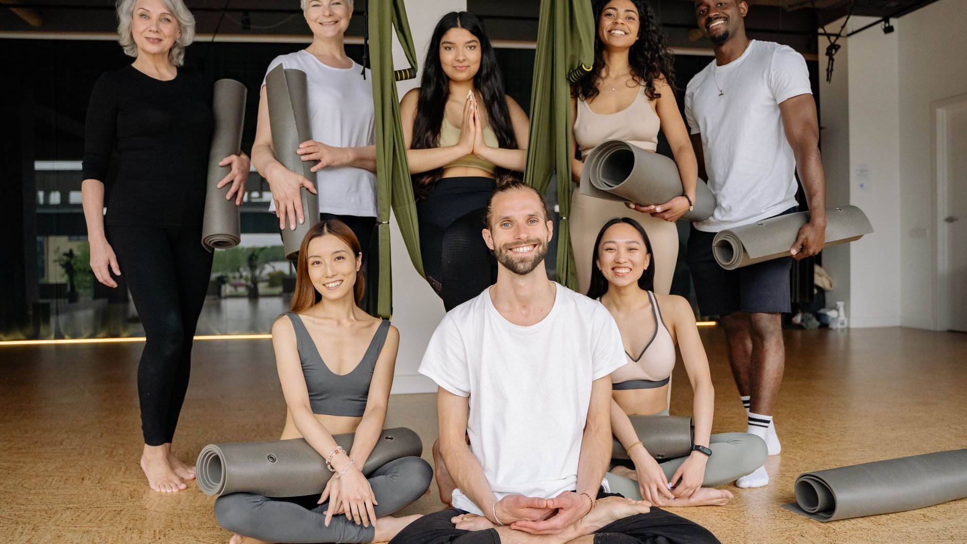 A group of smiling people with rolled yoga mats pose in a bright studio, some standing and some sitting cross-legged (illustrative stock photo)